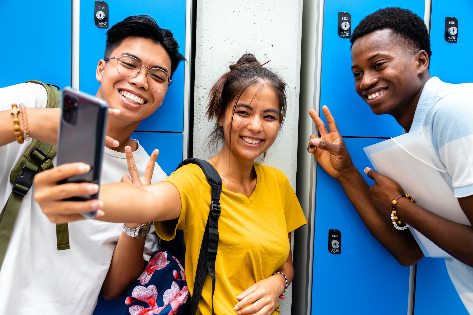 Smiling group of multiracial teen friends taking a selfie in high school corridor.